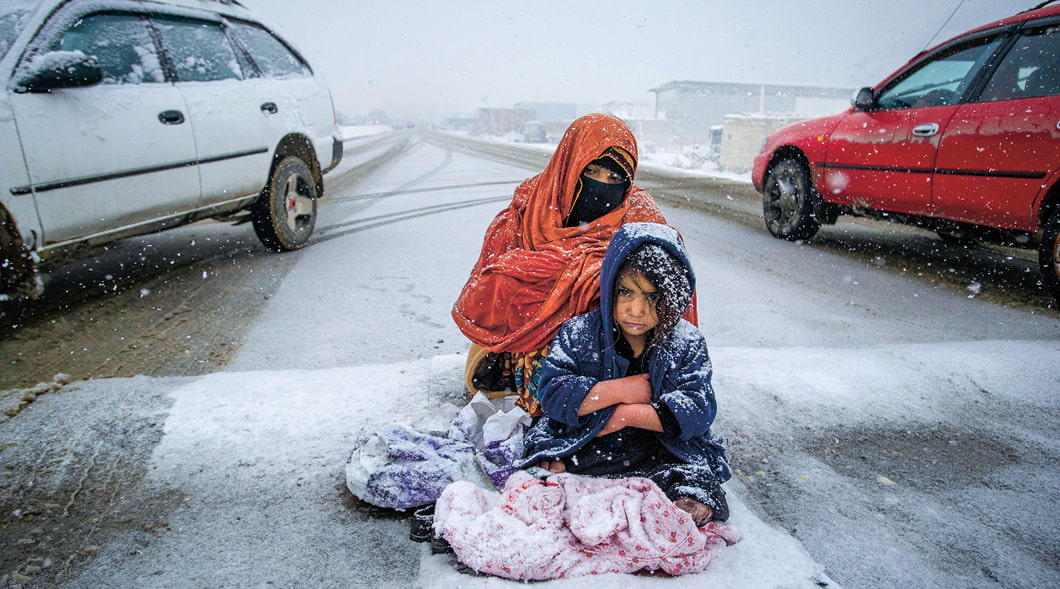 Two people sitting in the street between cars while it&apos;s snowing