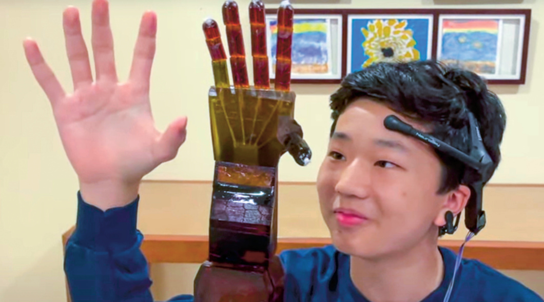 Person wearing a head piece while holding up hand next to a robotic hand