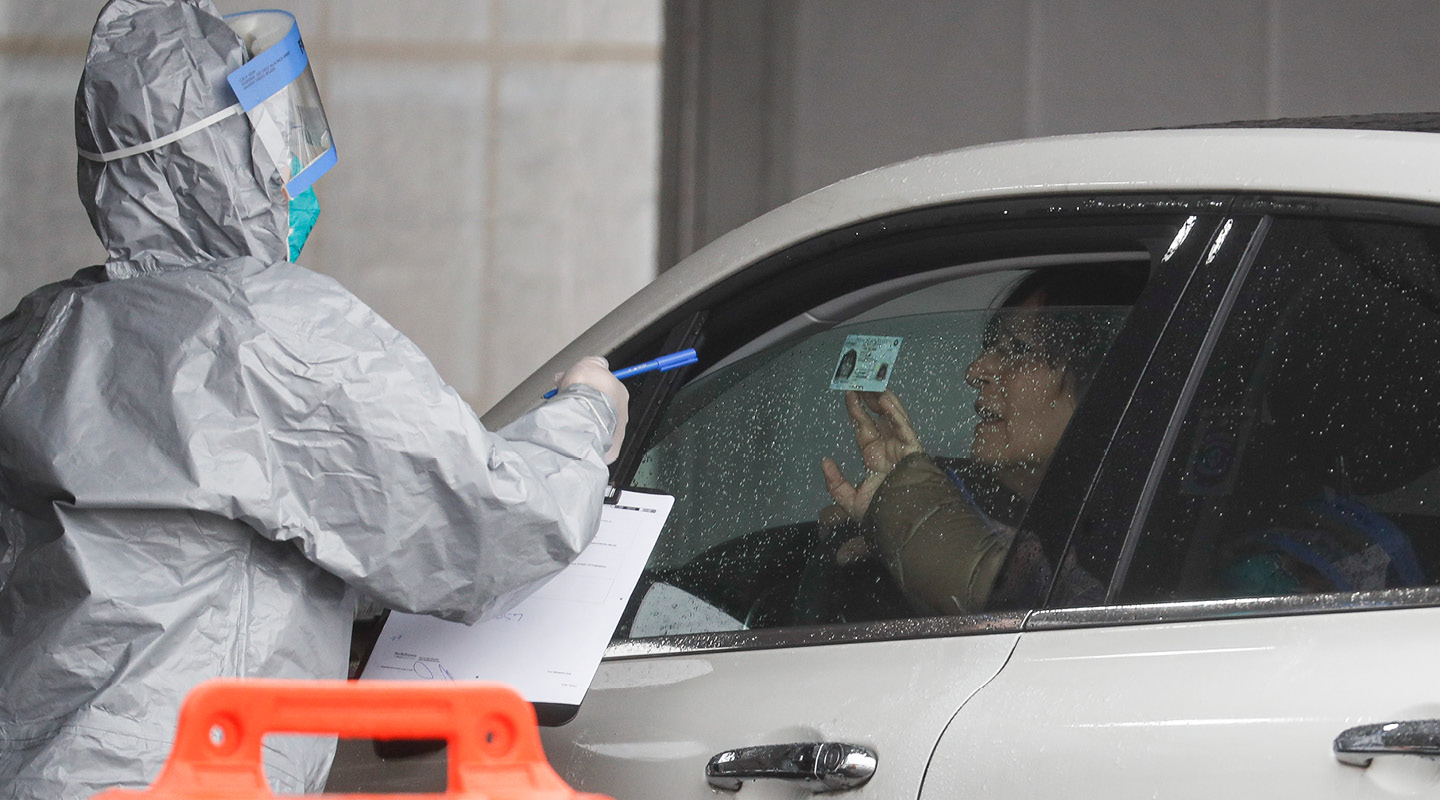 a man driving a car shows ID to a healthcare worker in a hazmat suit